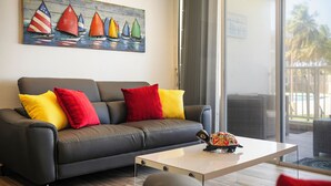 Ein modernes Wohnzimmer mit einem grauen Sofa, farbenfrohen Kissen, einem Gemälde von Segelbooten und Blick auf einen Balkon mit Palmen.