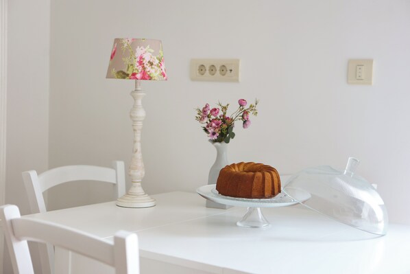 Una mesa con un pastel sobre un soporte de vidrio transparente, un florero con flores y una lámpara con pantalla floral.
