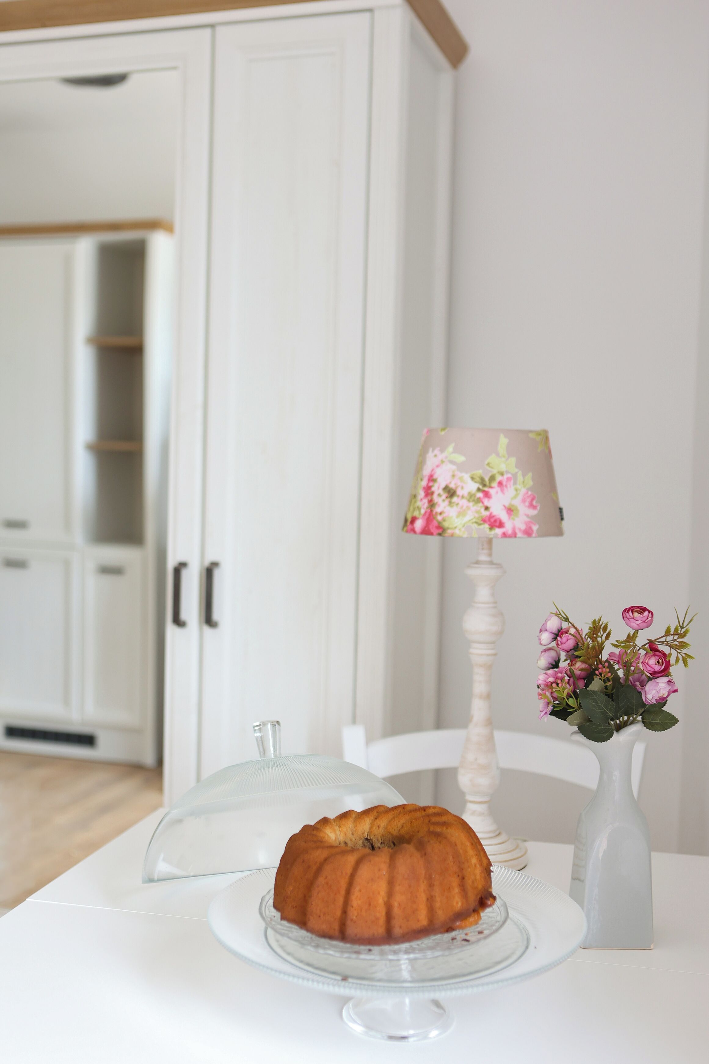 Una cocina con un pastel Bundt en un expositor de vidrio, una lámpara floral y un florero con flores sobre una mesa blanca.