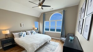 A bedroom with a bed, nightstands, a desk, a ceiling fan, and a view of the ocean through large windows.