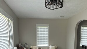 Een woonkamer met een hoekbank, twee fauteuils, een salontafel en een houten vloer. Er zijn twee ramen met zonwering, een plafondlamp en een kleed.