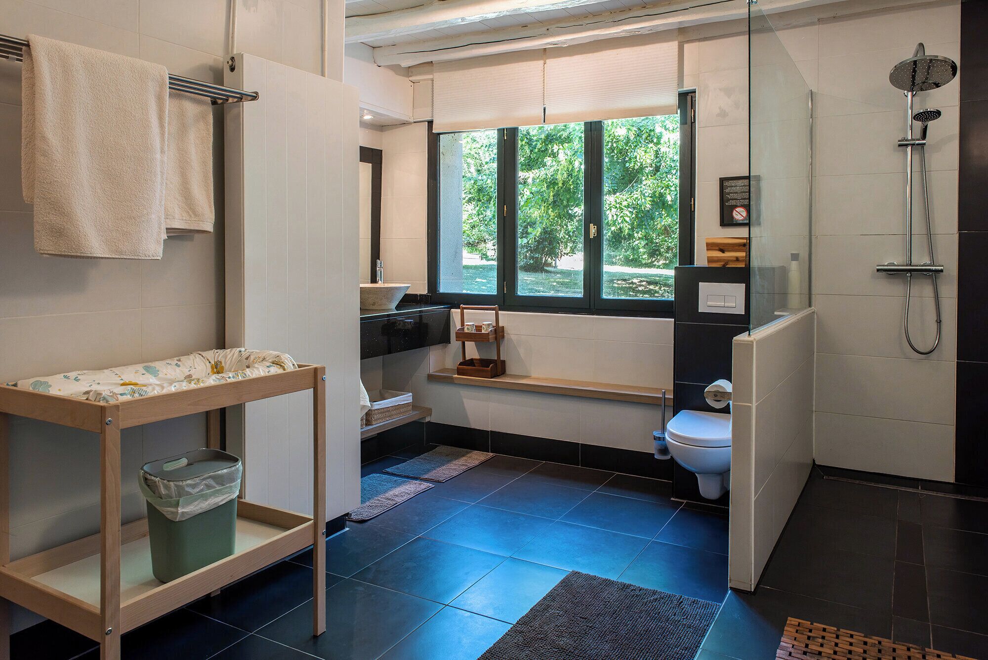 A modern bathroom with a bathtub, toilet, and sink. There is a window providing natural light and a view of greenery outside.