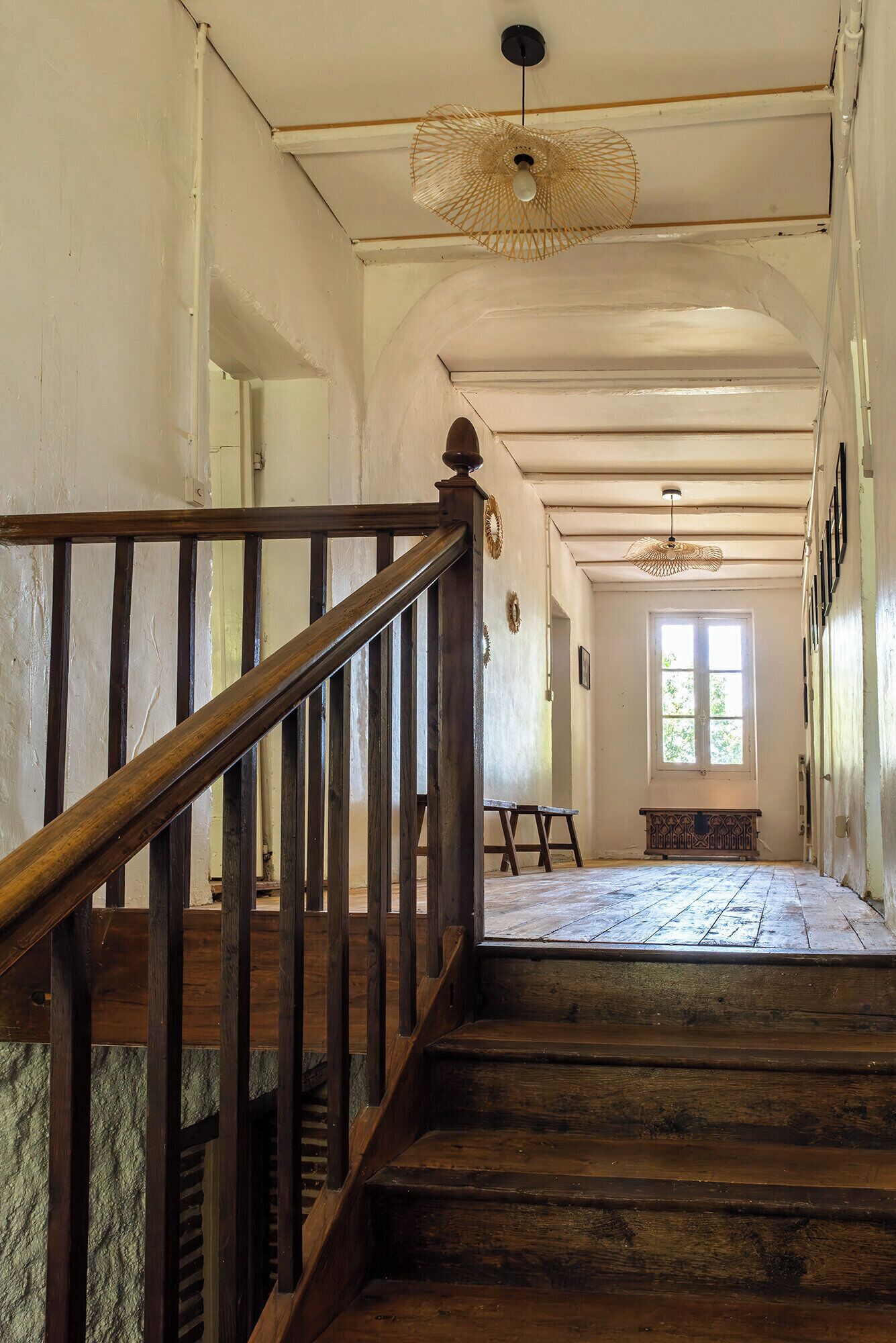 Un escalier en bois, doté d’une rampe, mène à un palier. Une fenêtre offre une vue sur la verdure environnante.