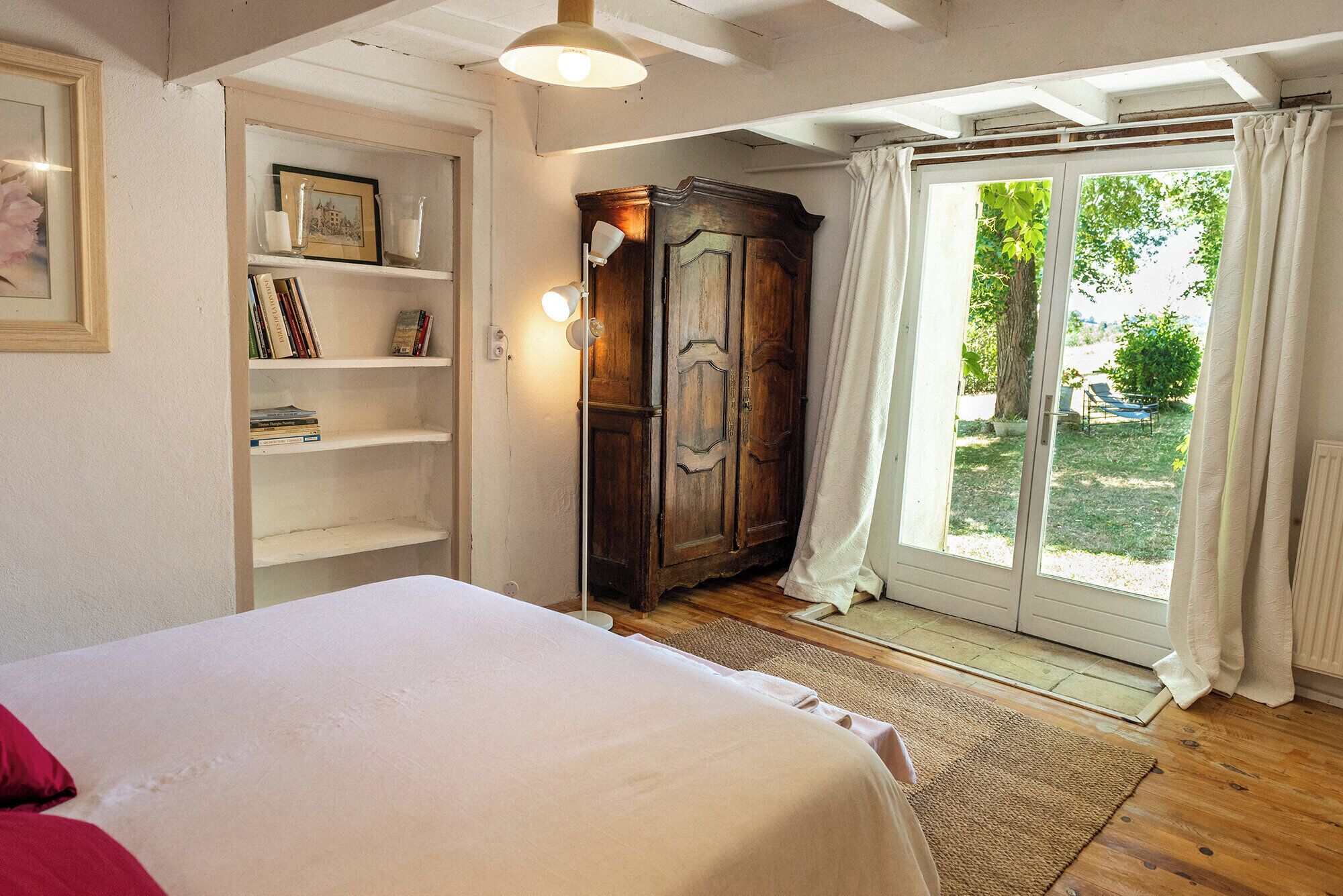 A bedroom with a bed, a wooden wardrobe, a bookshelf, and a sliding glass door to a garden.