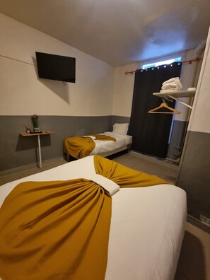 Une chambre d’hôtel avec deux lits, un téléviseur fixé au mur, une petite table avec un vase et une étagère avec des serviettes pliées.