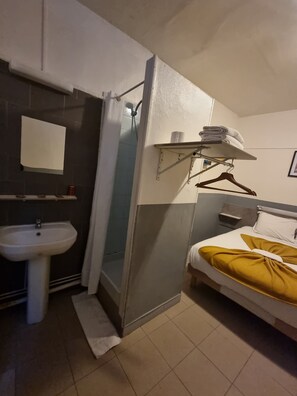 Une chambre d’hôtel de taille réduite, dotée d’une douche, d’un lavabo et d’un lit recouvert d’une couverture jaune.
