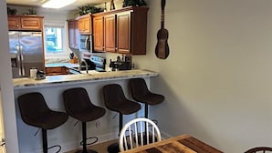 A kitchen with wooden cabinets, a refrigerator, a microwave, and a guitar on the wall. There are four bar stools and a dining table with a mug on it. - Riverfront AlpineRiverBear, Sleeps10, Walk To Oktoberfest! 2 HotTubs, HomeCinema (Helen)
