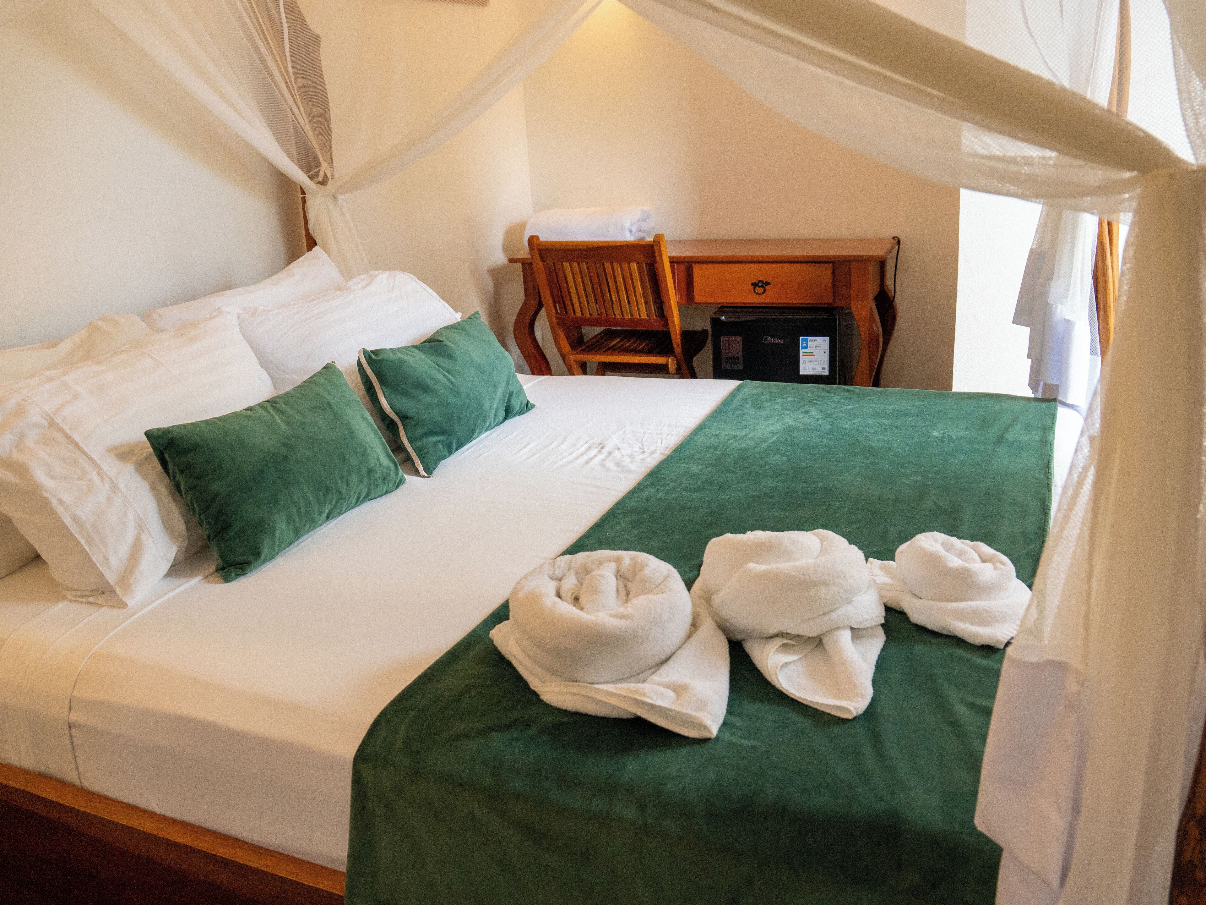 Una cama bien hecha con ropa de cama blanca y verde, una mesita de noche de madera y un pequeño escritorio con una silla.