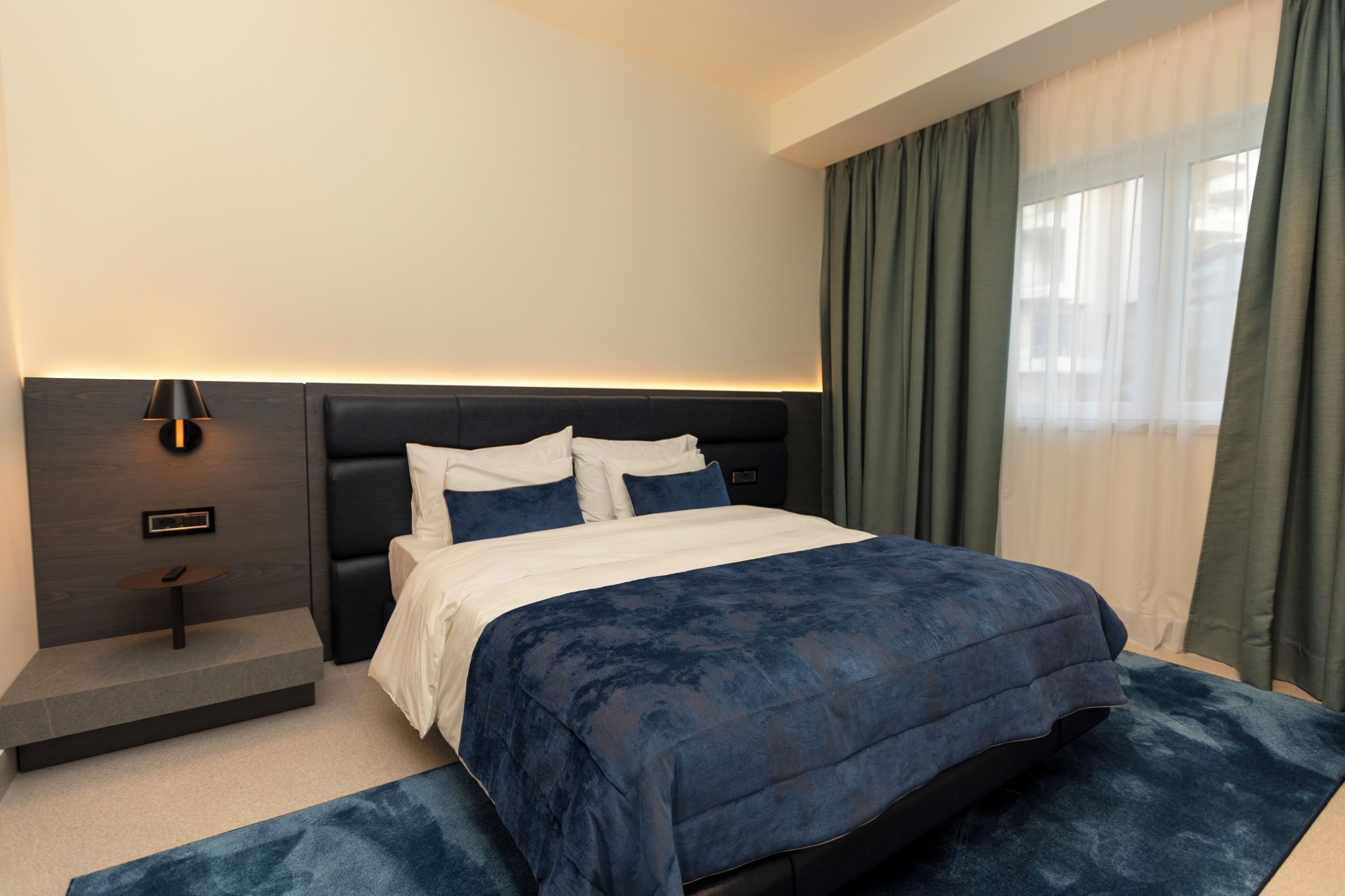A modern hotel room with a large bed, a bedside table, a lamp, and a view of the outdoors through the window.