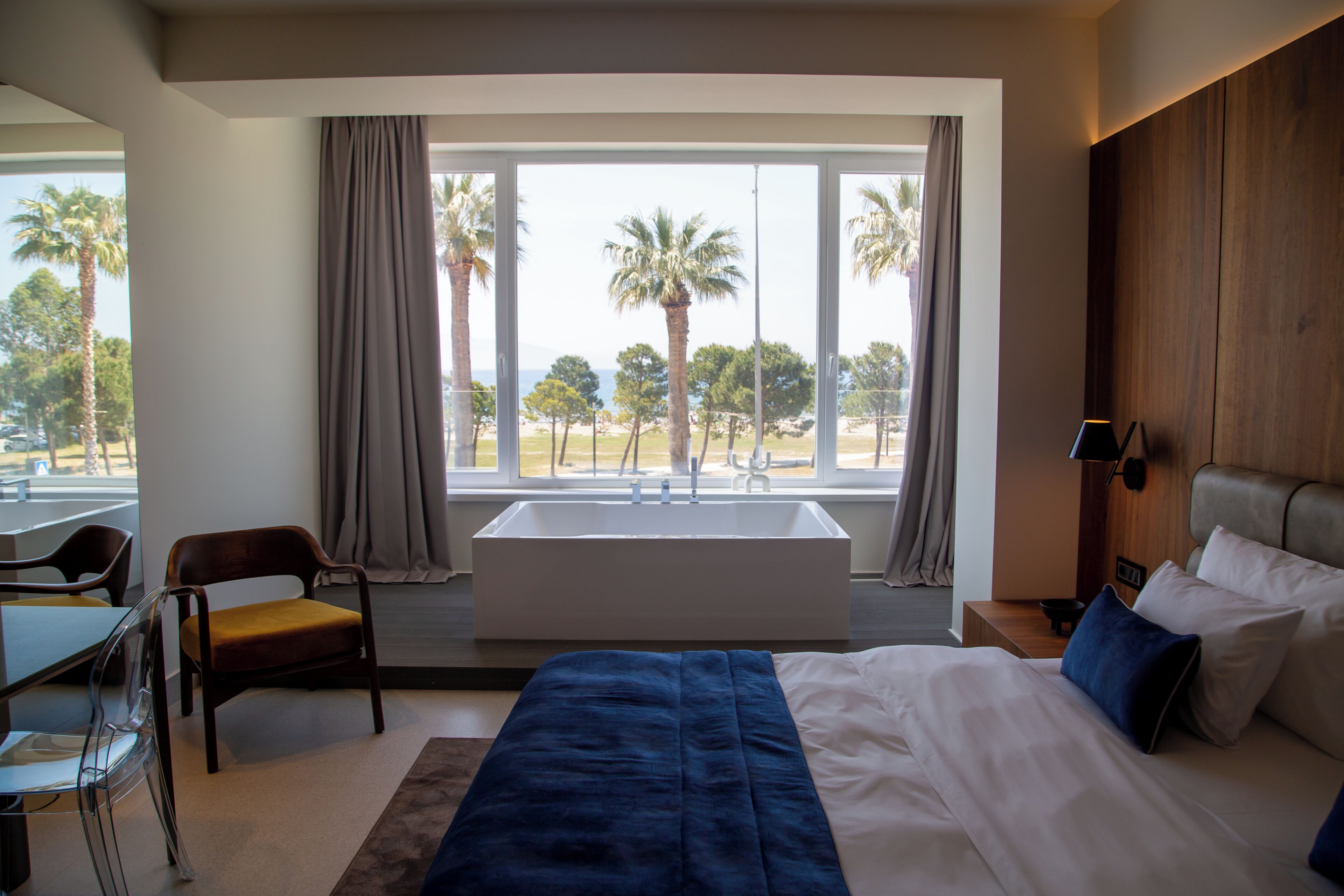 A hotel room with a large bed, a desk, a chair, a window with a view of palm trees and a beach, and a bathtub.