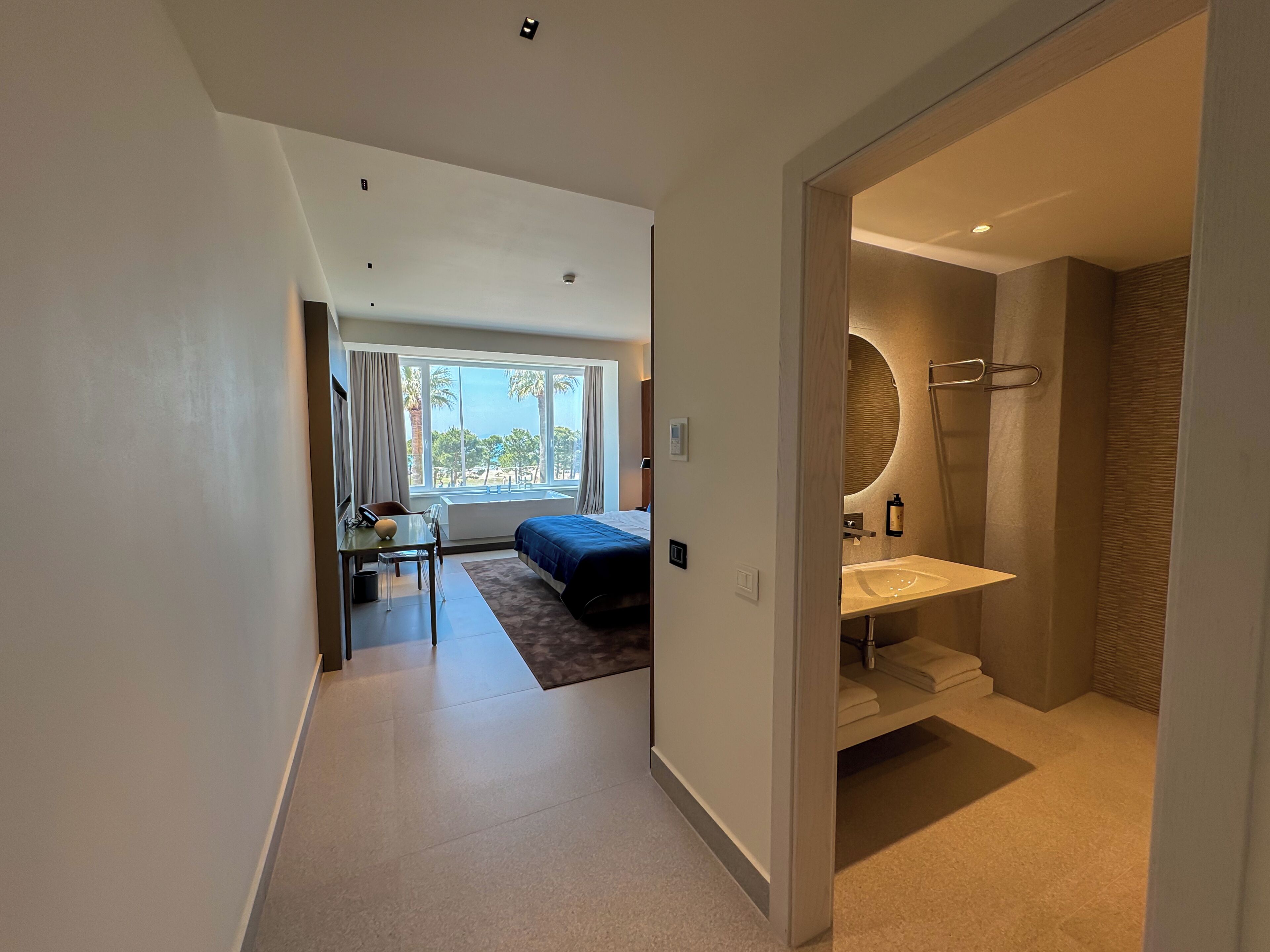 Ett modernt hotellrum med en säng, ett skrivbord, utsikt över stranden och ett badrum med handfat och spegel.