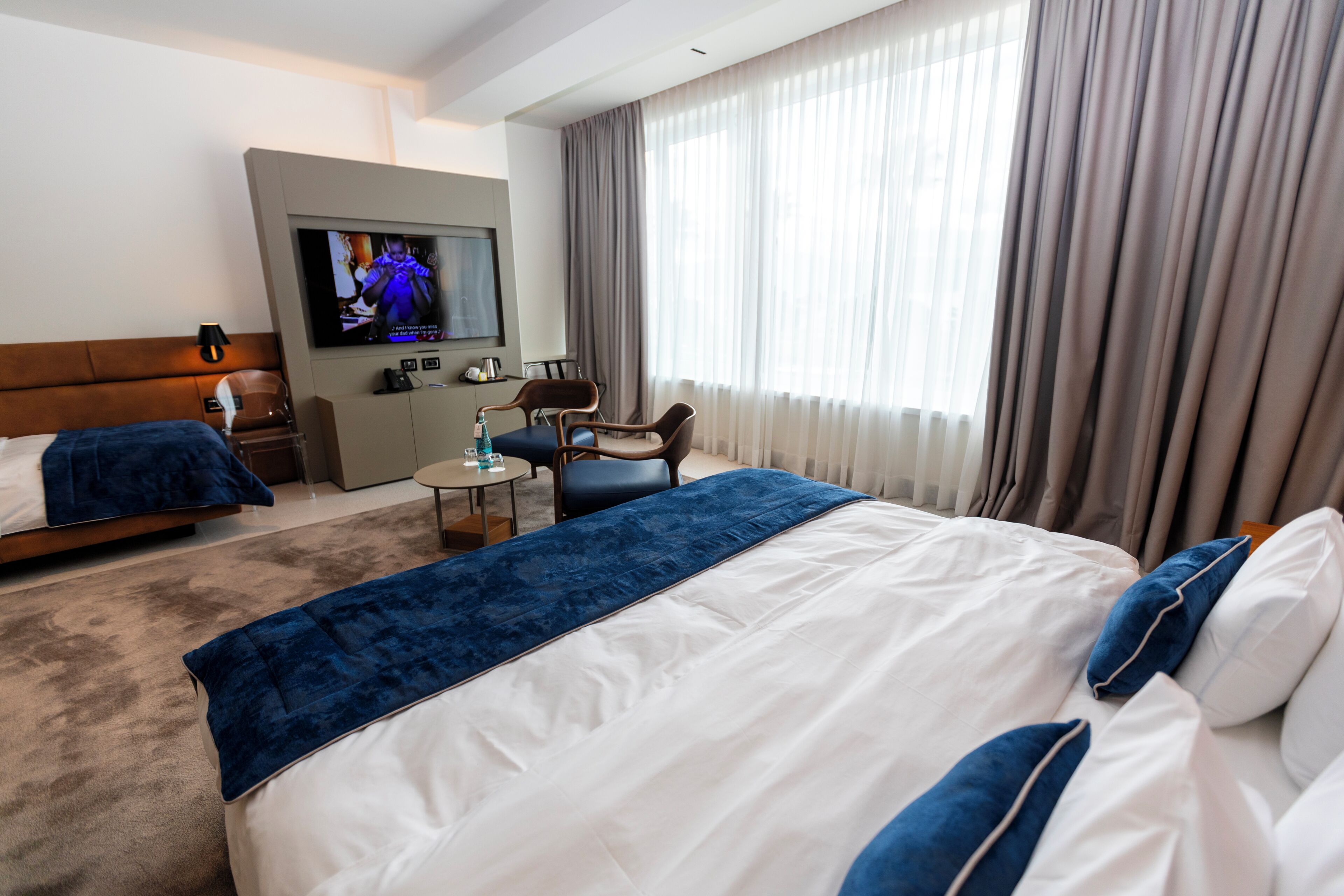 Habitación de hotel con una cama grande, una cama más pequeña, un escritorio, una silla, un televisor y una ventana con cortinas.