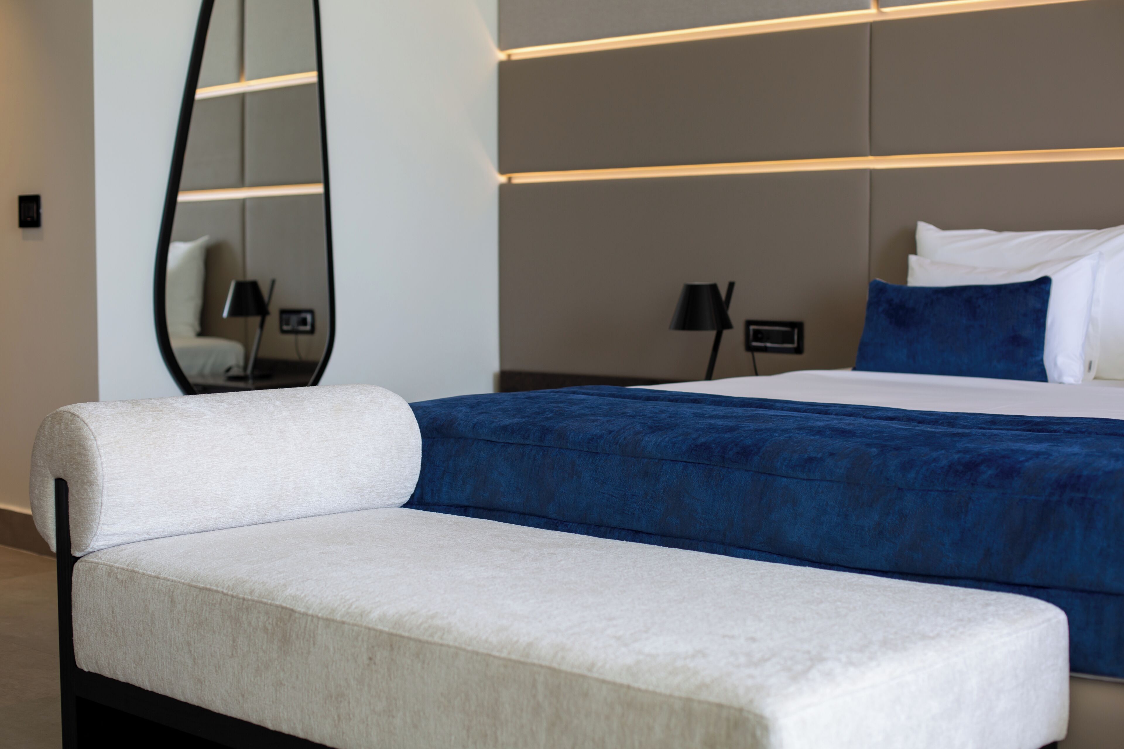 A modern hotel room with a large bed, a bedside table, a chair, a mirror, and a nightstand with a lamp.