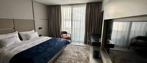 A modern hotel room with a large bed, a flat-screen TV, a seating area with a chair, and a large window with curtains.
