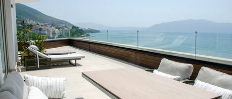 A balcony with a wooden table and white cushioned seating, overlooking a coastal cityscape.