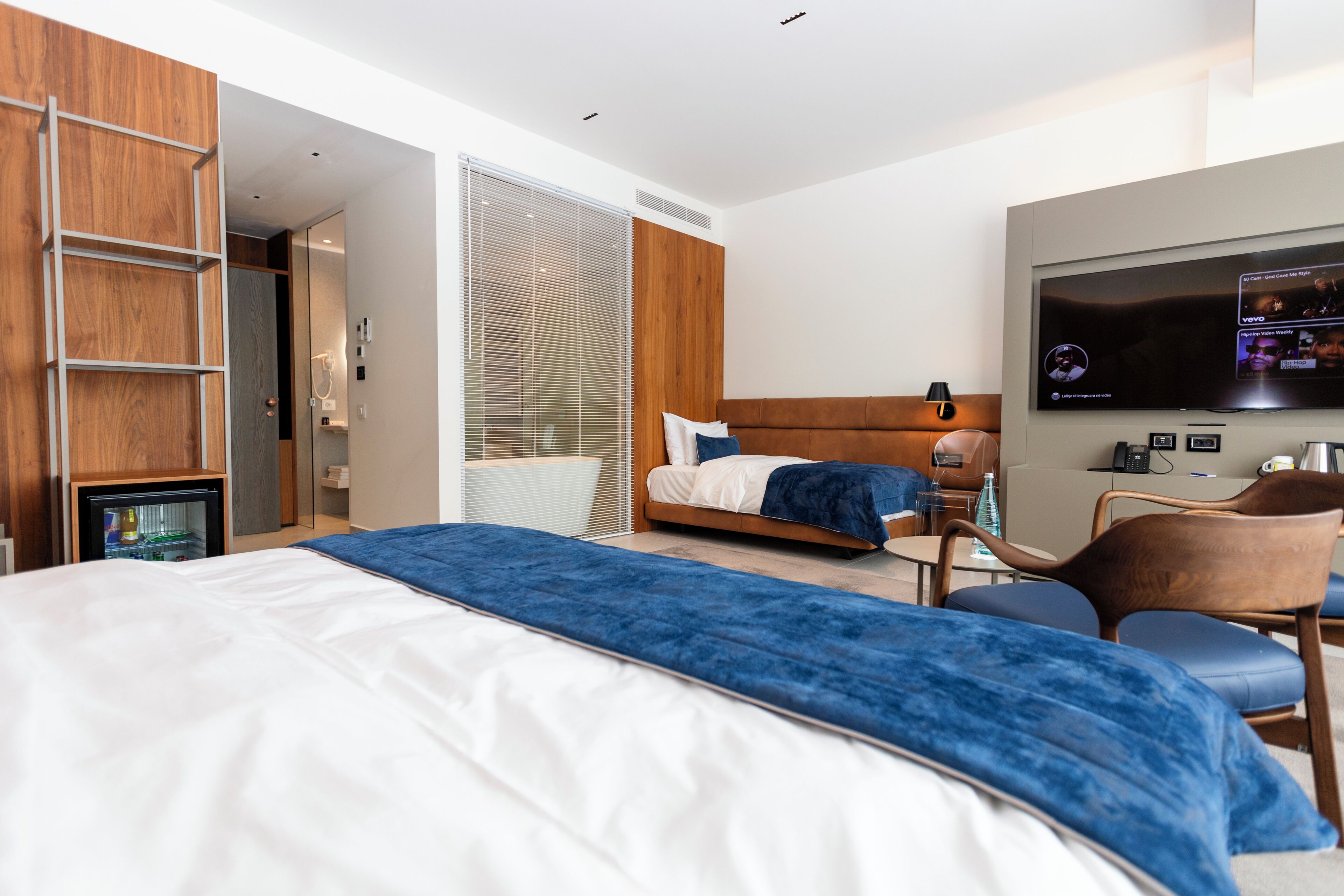 A modern hotel room with a large bed, a seating area with a chair, a television, and a minibar.
