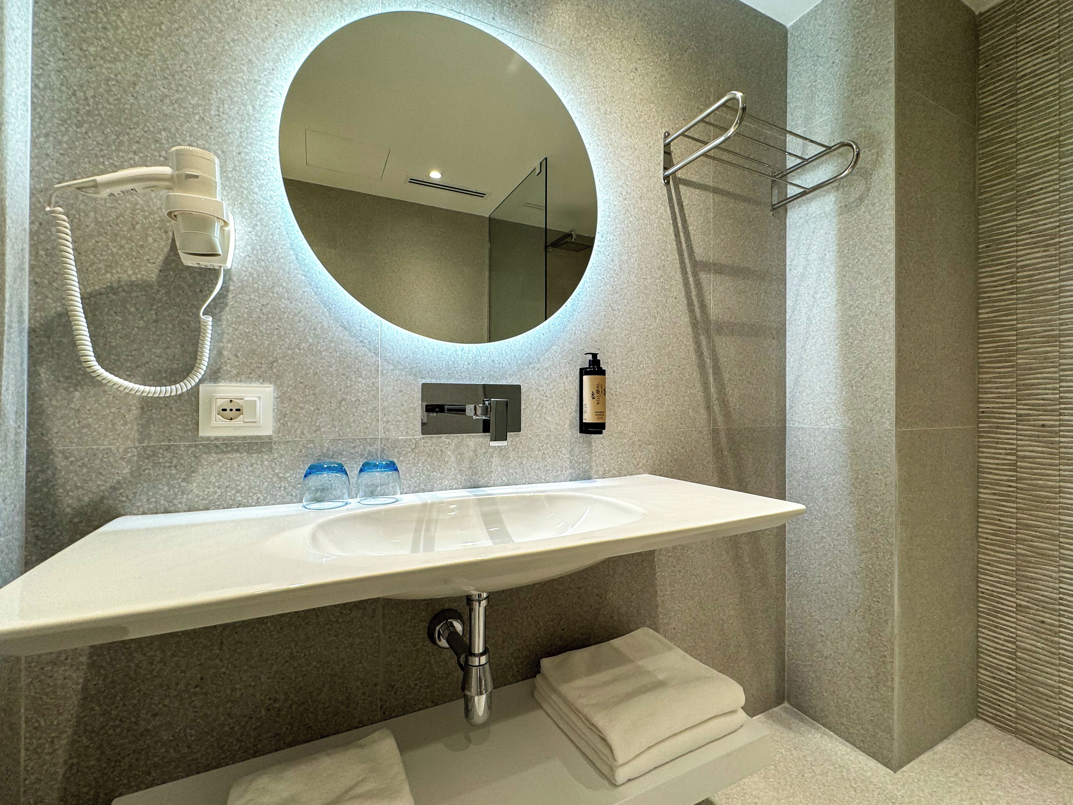 A modern bathroom with a white sink, a round mirror, a handheld showerhead, and a towel rack.