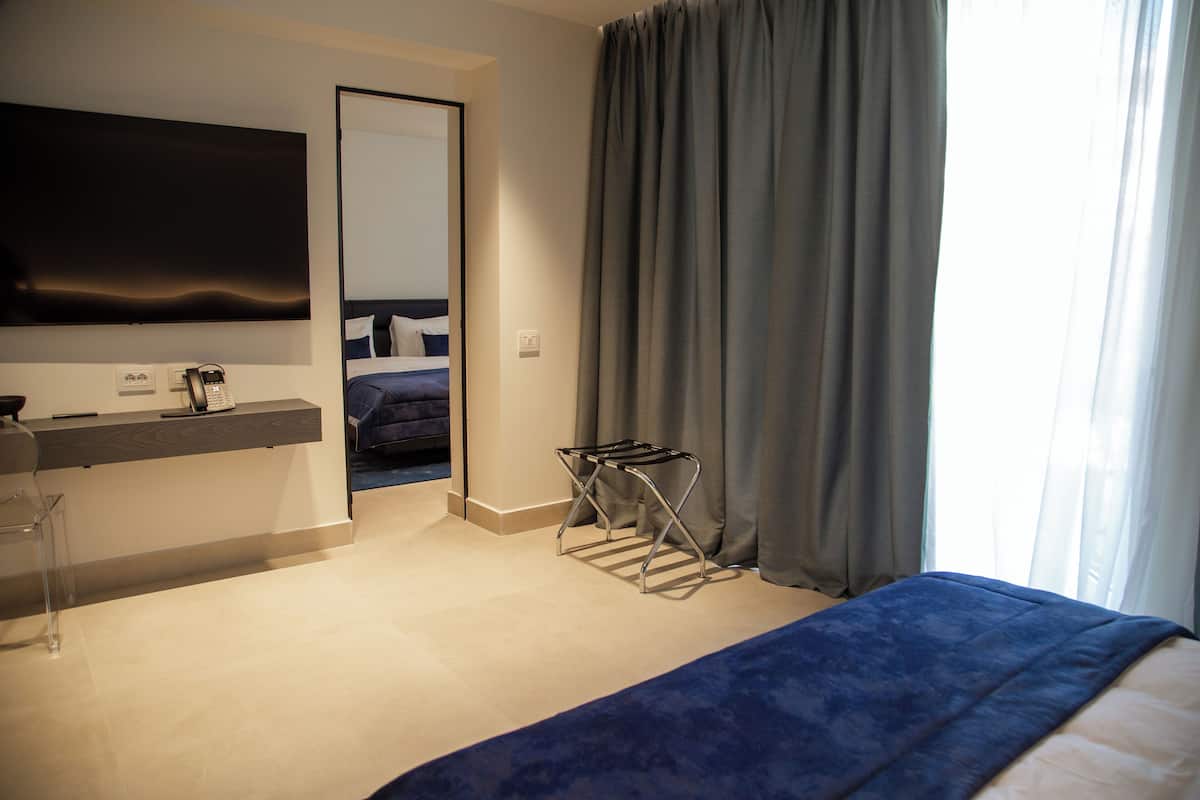 A modern hotel room with a bed, a flat-screen TV mounted on the wall, a desk with a telephone, and a window with curtains.
