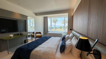 Habitación de hotel moderna con una cama grande, televisor de pantalla plana, escritorio con lámpara y vista a palmeras y al mar.