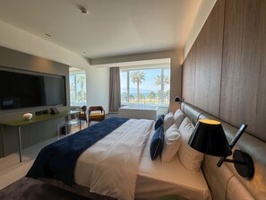 A modern hotel room with a large bed, a flat-screen TV, a desk with a lamp, and a view of palm trees and the sea. - Abi's Hotel & SPA Vlore (Vlorë)