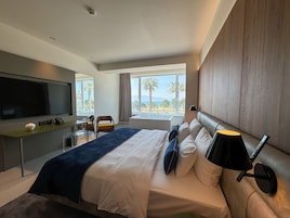 Quarto de hotel moderno com uma cama grande, uma TV de tela plana, uma escrivaninha com abajur e vista para palmeiras e o mar.