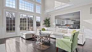 A spacious living room with a large window, a dining area, and a kitchen visible through an open doorway.