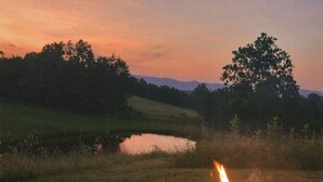Una fogata en un brasero metálico, con un campo de hierba y un cuerpo de agua al fondo durante el atardecer.