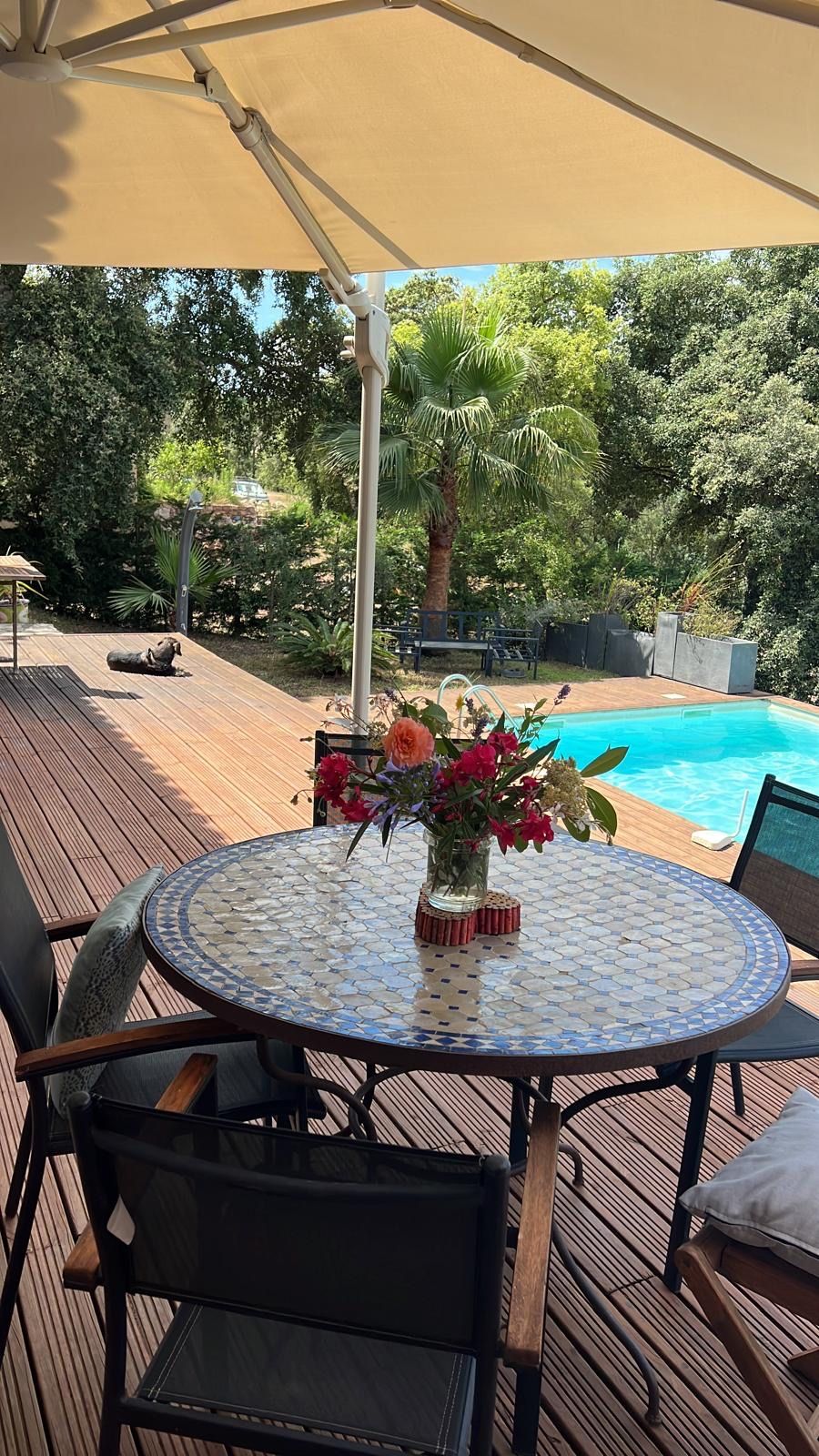 A round table with a mosaic top, surrounded by chairs, set outdoors with a pool and greenery in the background.