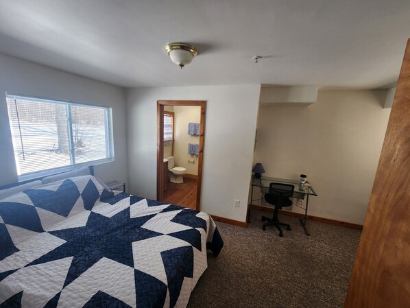 A bedroom with a large bed, a desk with a chair, a window with blinds, and a door leading to a bathroom.