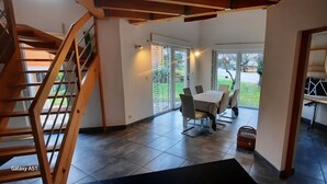 Amplia habitación con escalera de madera, zona de comedor con mesa y sillas, y vistas al exterior a través de amplios ventanales.