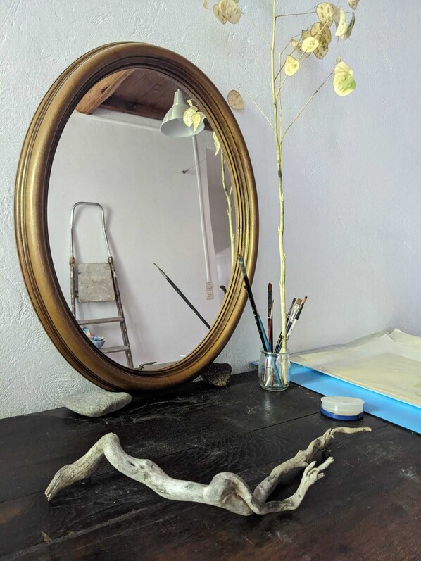 A mirror with a gold frame on a dark wooden surface, a vase with dried flowers, and a small container with paintbrushes. - House on the river (Ganges)