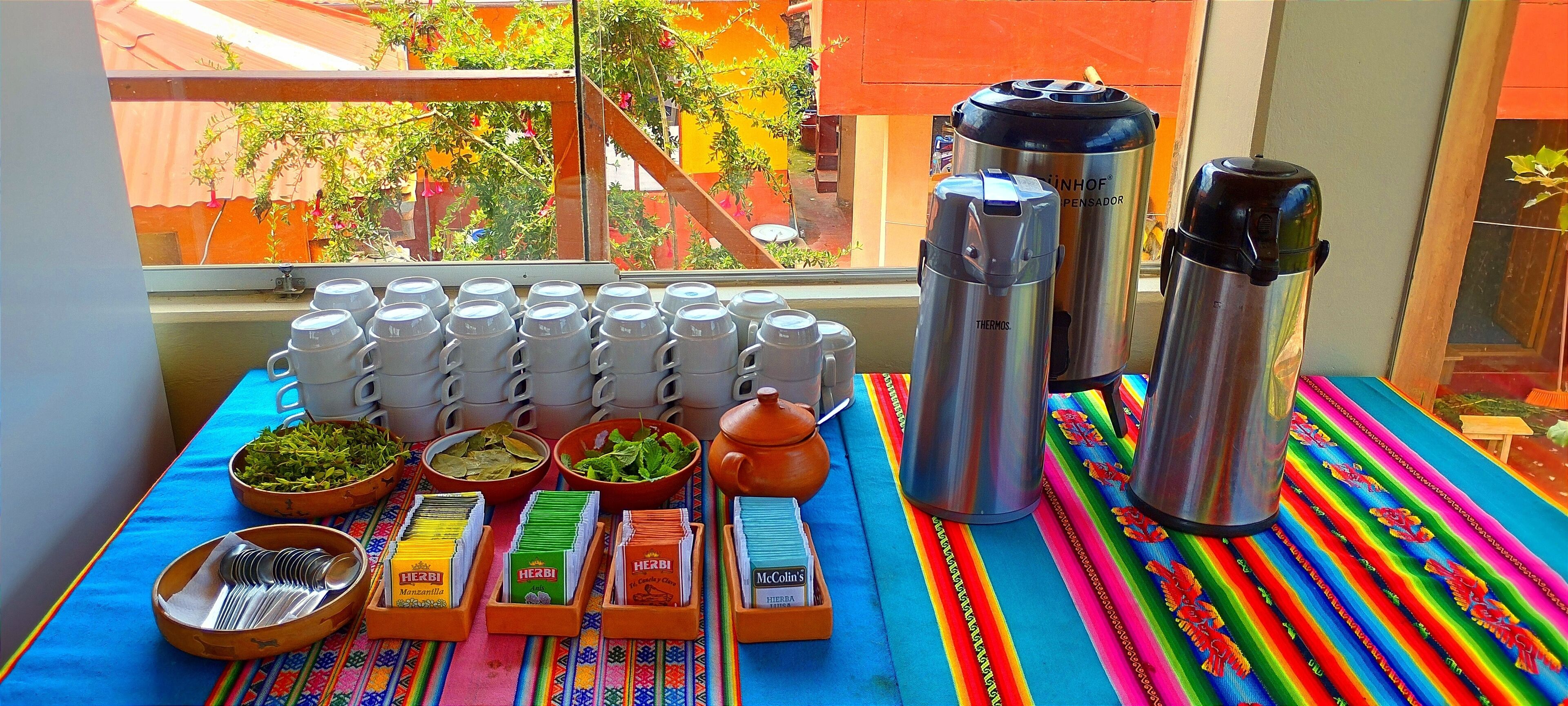 Una mesa con termos, tazas y aderezos, sobre un mantel colorido, con vista a un edificio.