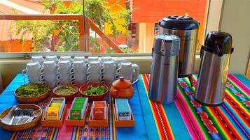 Una mesa con termos, tazas y aderezos, sobre un mantel colorido, con vista a un edificio.