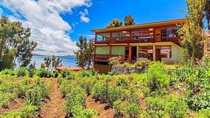 Un edificio de dos plantas con balcón, rodeado de follaje y vista al mar.