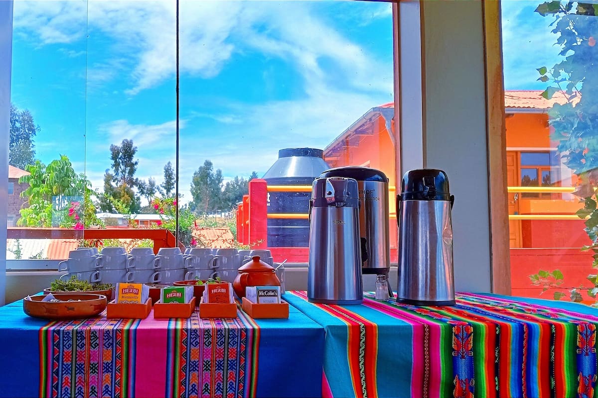 Une table avec des thermos et des récipients en céramique, une nappe colorée, et une vue sur un bâtiment rouge et des arbres, visible à travers une fenêtre.