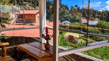 Mesa e banco de madeira dentro de um quarto com uma janela grande para um monte com casas e vegetação.