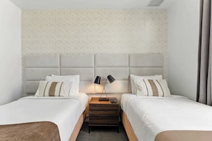 Habitación de hotel con dos camas, cabecera con diseño geométrico, mesitas de noche con lámparas y un cajón pequeño.