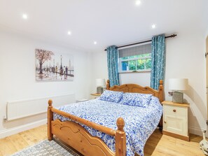 A bedroom with a wooden bed, a nightstand with a lamp, a window with curtains, a radiator, and a wall-mounted picture.