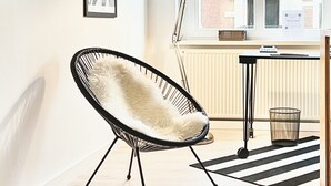 Ett rum med ett stort fönster, en svart stol, ett bord med en lampa och inramade tavlor på väggen.