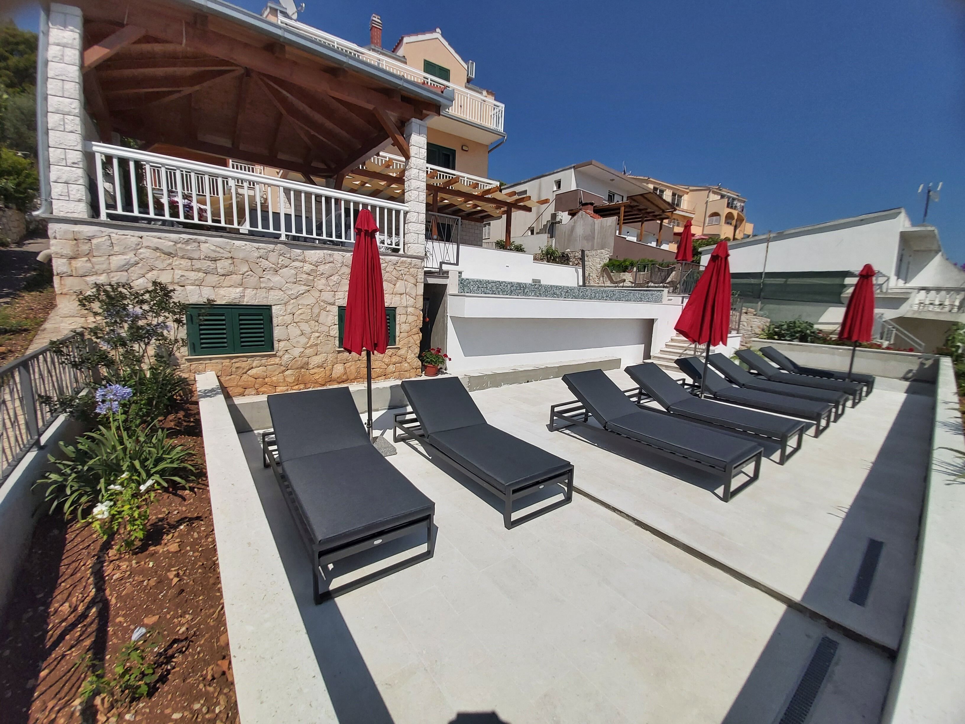 A rooftop terrace with sun loungers and red umbrellas, stone walls, and a clear blue sky.