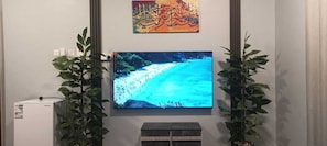 A television mounted on a wall displaying a beach scene, flanked by two potted plants. - Al Ghazali Suites (Madinah)