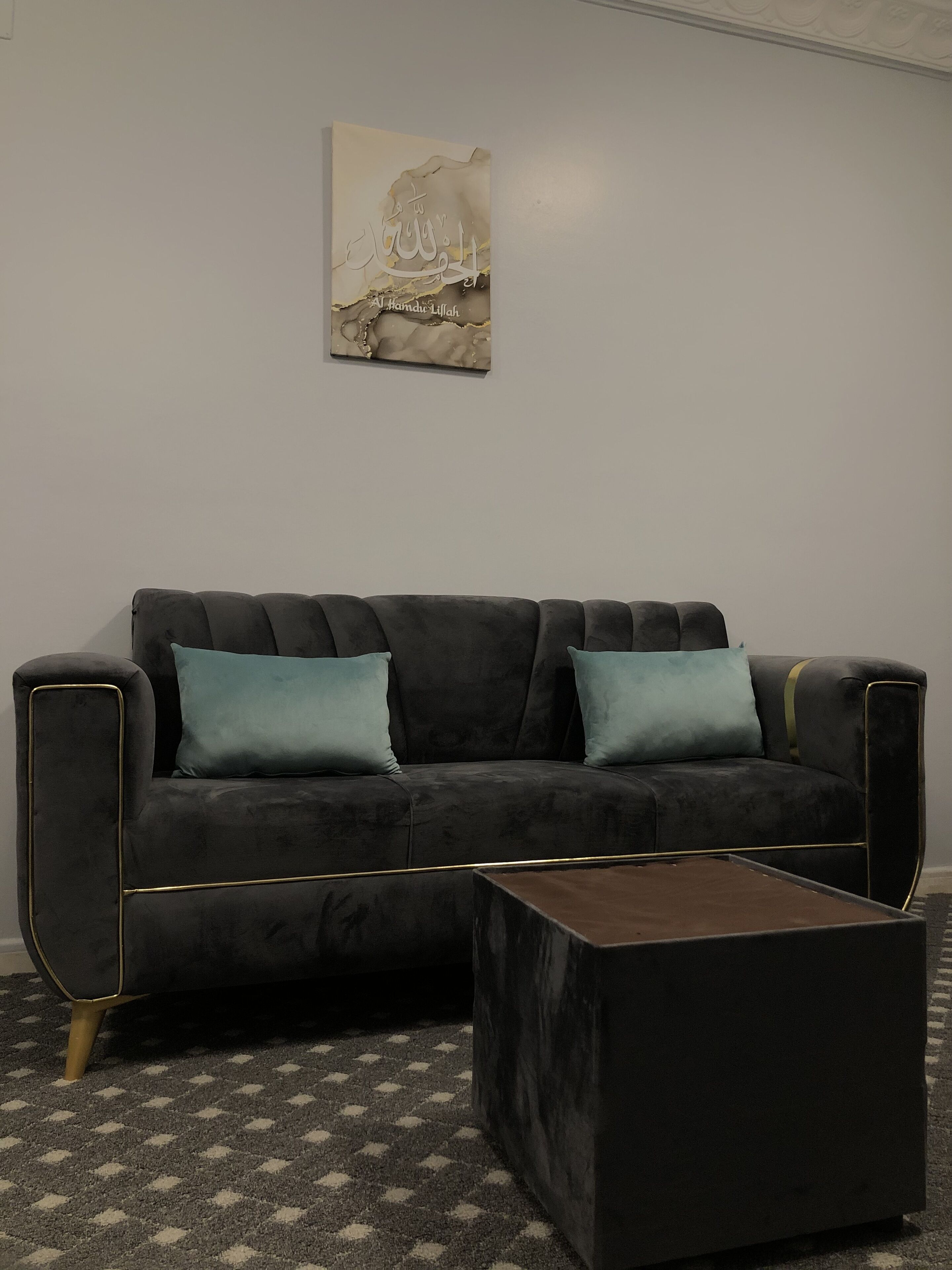 Una sala moderna con un sofá gris oscuro, dos cojines color turquesa y una pequeña mesa de centro de madera. Una obra de arte enmarcada cuelga en la pared blanca lisa.