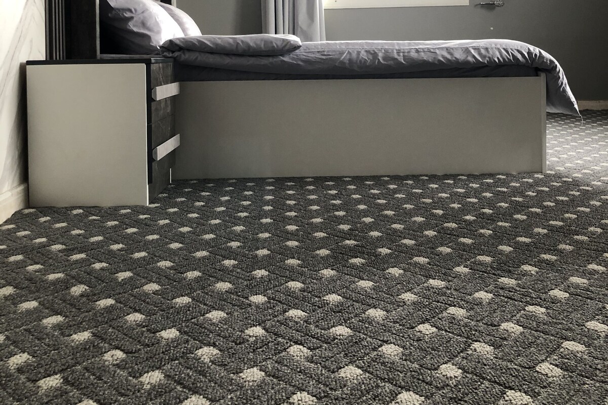 A modern bedroom with a bed, a nightstand, a window with curtains, and a patterned carpet.