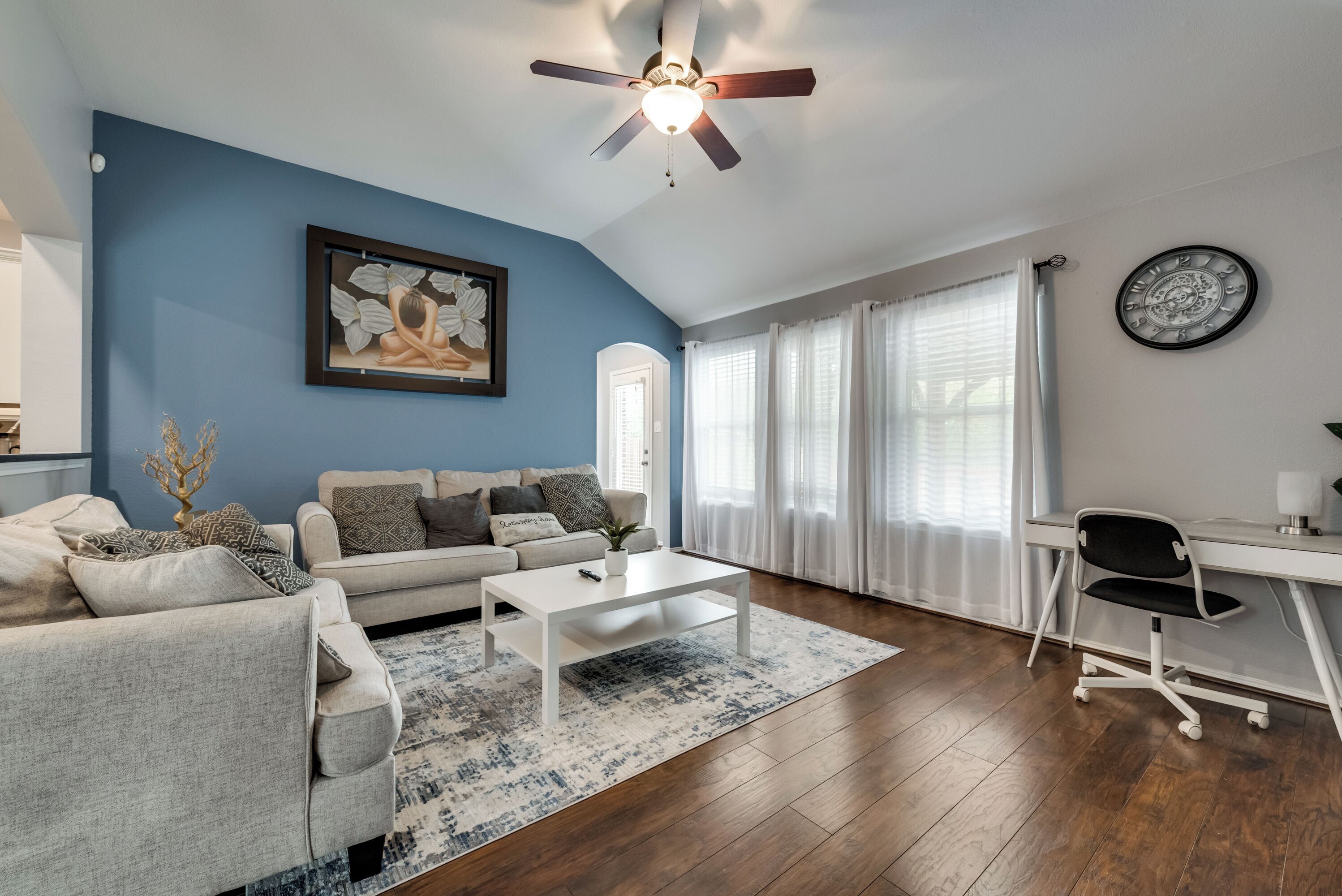 Una sala de estar con una pared azul decorativa, un ventilador de techo, un cuadro, un sofá con cojines estampados, una mesa de centro, una alfombra, un escritorio con una silla y un reloj en la pared.