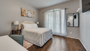 A bedroom with a bed, a desk, a chair, a painting, a window with curtains, and a mirror.