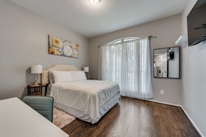 Un dormitorio con cama, escritorio, silla, un cuadro, ventana con cortinas y espejo.