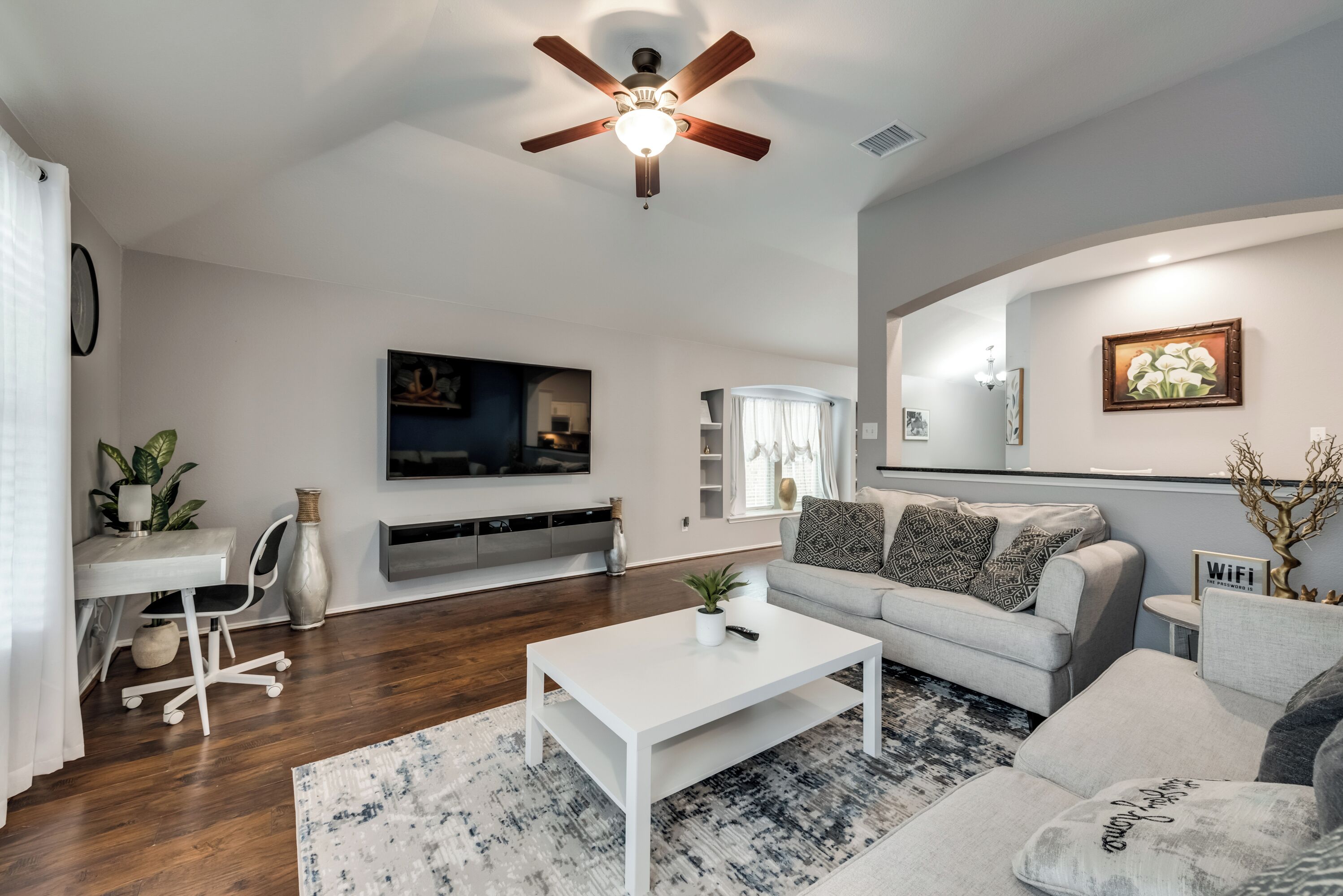 A modern living room with a ceiling fan, a flat-screen TV mounted on the wall, a grey sofa with patterned cushions, a white coffee table, and a dining area with a white table and chairs.