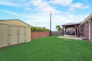 A backyard with a shed, a covered patio area, and a brick wall. - GP020 | G. Prairie Home | 3BD | Yard | Garage (Grand Prairie)