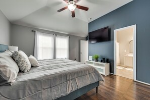 Un dormitorio con una cama, un ventilador de techo, un televisor en la pared y un baño visible a través de una puerta abierta.