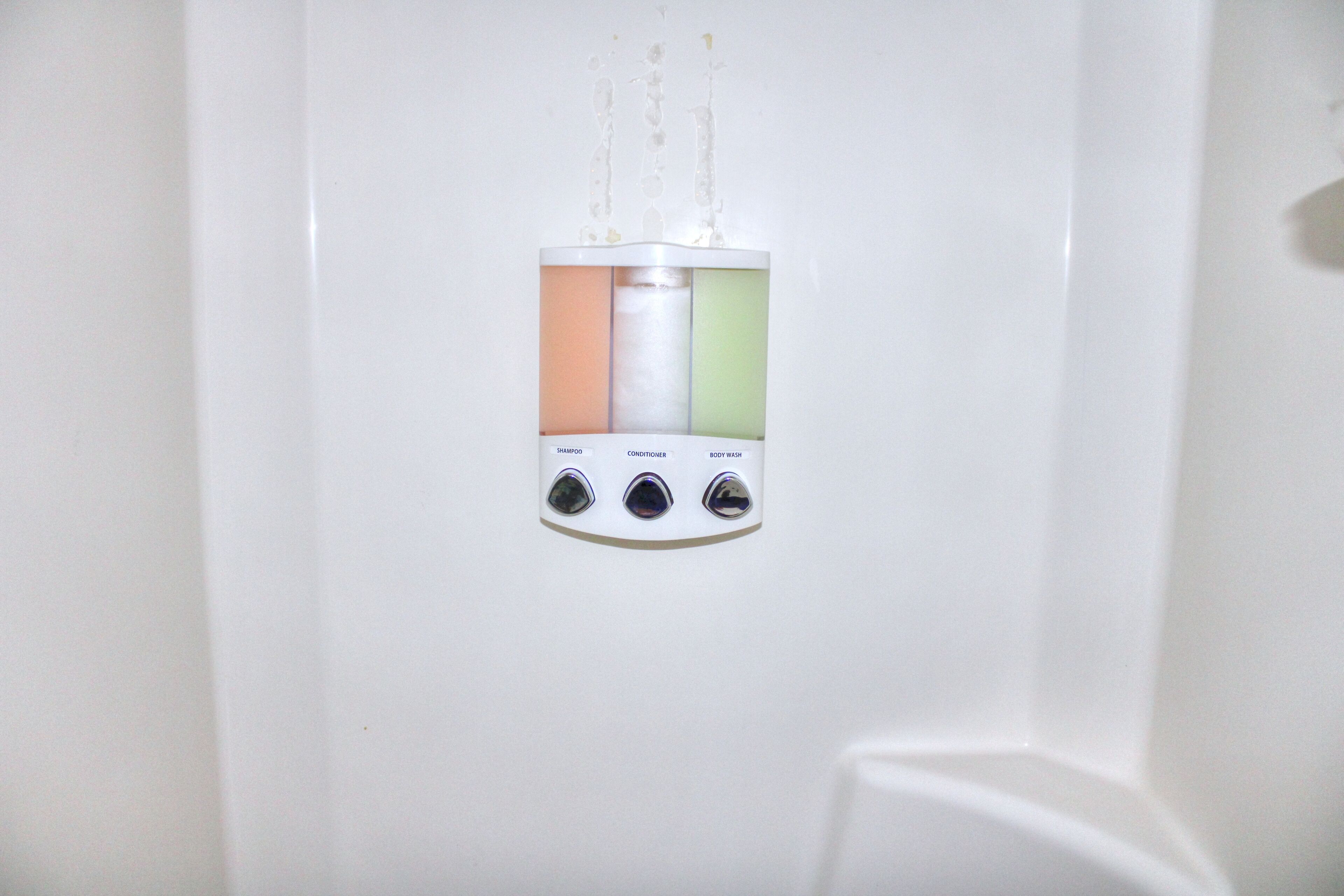 A wall-mounted dispenser with three compartments labeled for shampoo, conditioner, and body wash.
